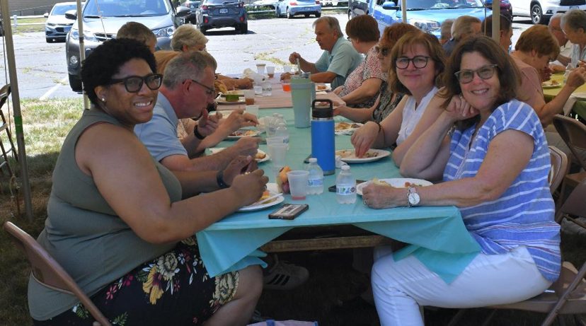 Church members smiling at a long table under a tent at a summer lunch