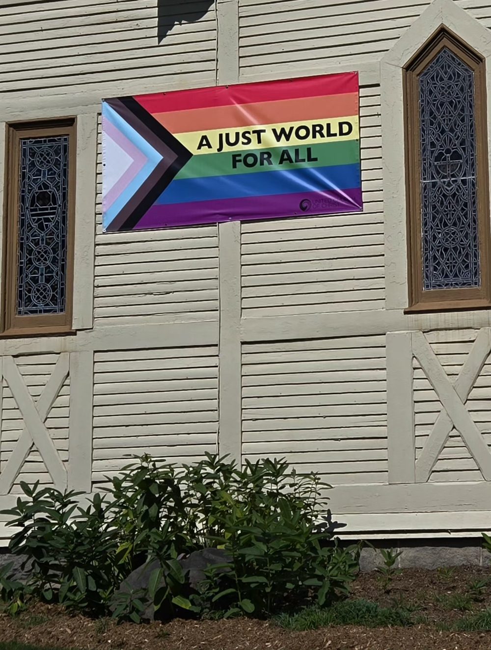 A Progress Pride banner reading “A Just World for All” hanging on the church’s wooden siding between two stained glass windows